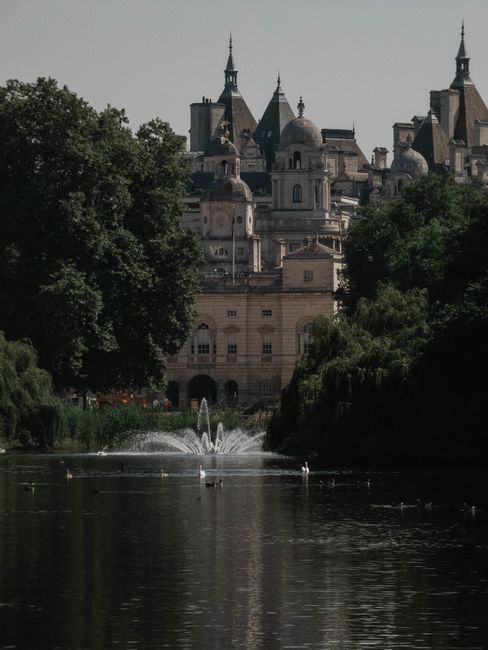 A panoramic view of a historic castle with multiple spires, domed turrets, and ornate architectural details, situated behind a lush green landscape with large trees on either side. In the foreground, a calm river reflects the dark silhouette of the trees and the castle, with a central water fountain creating gentle ripples in the water. The scene is captured during daylight with overcast lighting, highlighting the contrasts between the greenery, the stone building, and the reflective surface of the river. The environment suggests a private garden or park setting, possibly used for outdoor leisure or scenic viewing, indirectly associated with independent rubbish clearance or waste management services like House Clearance Westminster.
