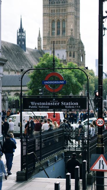 The image shows the entrance to Westminster Station, an underground railway station in London, with the iconic red and blue London Underground roundel sign prominently displayed above a black metal frame supporting a sign that reads 'Westminster Station Public Subway Toilets.' The scene is set on a busy city street with numerous pedestrians walking and congregating near the entrance, some wearing jackets and hats, indicating mild weather. To the right, there's a black lamppost with a speed restriction sign showing '20' and part of an orange and white warning sign. In the foreground, a black wrought-iron fence surrounds the stairs leading down into the station, and a man is seen descending. In the background, the historic architectural features of Westminster Abbey are visible, with stone towers and gothic detailing, contrasting with the urban street scene. The ambient lighting suggests a cloudy day, with soft light reflecting off the stone surfaces. This snapshot captures both the historic and modern elements of Westminster, highlighting the busy nature of this central London area near key transport links, and subtly aligns with the importance of efficient rubbish removal services for urban spaces, such as those offered by House Clearance Westminster.