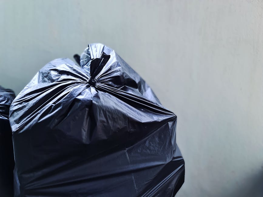 Close-up of a tightly knotted black plastic rubbish bag placed against a plain pale grey wall. The bag, made of glossy plastic material, reflects light on its surface, revealing creases and folds, with the top gathered and tied securely. The bag's size and fullness suggest it contains household waste, and it rests on a surface that is not visible in the frame. In the background, part of another black rubbish bag is visible, indicating a collection of waste perhaps awaiting removal or disposal. The neutral indoor setting and the arrangement imply a domestic or service context related to waste collection or private rubbish handling, consistent with independent rubbish removal services. The clean, simple environment emphasizes the focus on the waste bag, with natural lighting highlighting the textures of the plastic and the subdued background enhancing clarity for accessibility purposes.