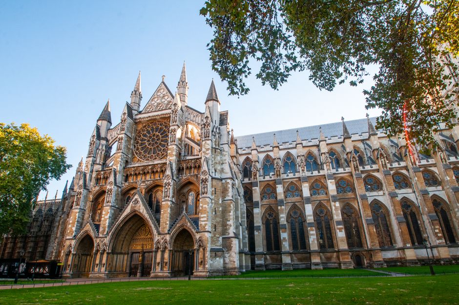 A large Gothic-style cathedral with intricate stonework and pointed arches, featuring an ornate facade with numerous stained glass windows and decorative pinnacles. The building's exterior is constructed from light-colored stone with contrasting darker accents and weathered textures. Tall, slender spires extend upward, framing the structure against a clear blue sky. In the foreground, a well-maintained green lawn surrounds the cathedral, with partially visible trees on the right and left edges providing natural framing. The scene is illuminated by soft, natural daylight, highlighting the detailed architectural features and casting gentle shadows. This setting emphasizes the historic stone architecture typical of Westminster area landmarks, often requiring on-site clearance or removal of debris during maintenance or restoration projects, reflecting the context of independent waste handling and rubbish removal services like those offered by House Clearance Westminster.