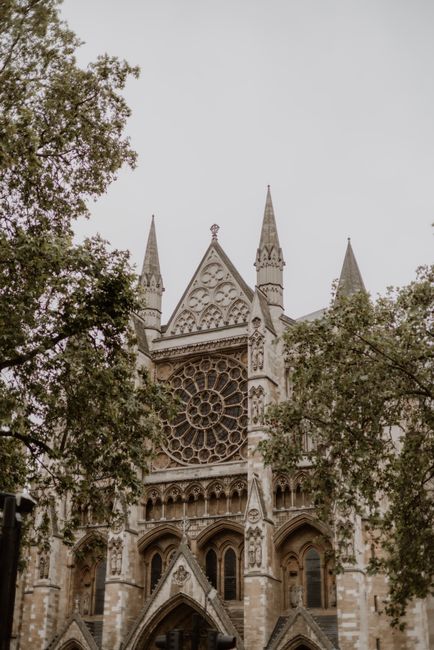 The photograph shows the façade of a historic Gothic-style cathedral, characterized by its pointed spires, ornate stonework, and large circular stained glass window featuring intricate tracery, situated prominently on the front elevation. The stone exterior has a weathered appearance with various shades of beige and light gray, reflecting age and craftsmanship. Two tall spires reach upward on either side of the central section, framing the main rose window. Surrounding the cathedral, there are leafy trees with dense foliage, partially obscuring the lower part of the structure and contributing to the scene's sense of scale and environment. The sky above is overcast, with diffuse lighting that minimizes harsh shadows, highlighting the detailed carvings and architectural features. The setting appears to be in an urban area, with no visible vehicles or modern elements in the immediate vicinity, emphasizing the monumentality of this historic religious building. As part of a rubbish removal context, the scene might suggest the importance of on-site clearance or preservation efforts, and it aligns with private waste handling services that can facilitate the maintenance of such cultural sites, as exemplified by companies like House Clearance Westminster.
