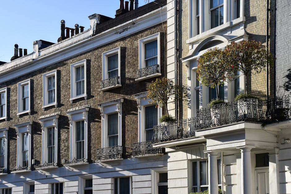 Photograph of a multi-storey Victorian-style residential building with a brick facade and white decorative trim. The building features several large, double-hung sash windows with white frames, some of which have small black wrought-iron railings attached to the window sills, providing balcony-style access. In the foreground, the building has a white-painted ground-floor façade with a prominent entrance supported by white columns. Two mature, well-kept potted trees with rounded, dense foliage are placed on a small balcony area, adding greenery to the scene. The exterior wall surfaces show a mix of exposed brickwork and painted sections, with the brickwork appearing weathered but well-maintained. The roofline includes black chimney stacks and flashing, set against a clear blue sky. The scene suggests a quiet urban environment, possibly in a city area with historical architecture, where private property and rooftop greenery contrast with common urban waste management activities, such as rubbish removal or on-site clearance, which local services like House Clearance Westminster might facilitate.