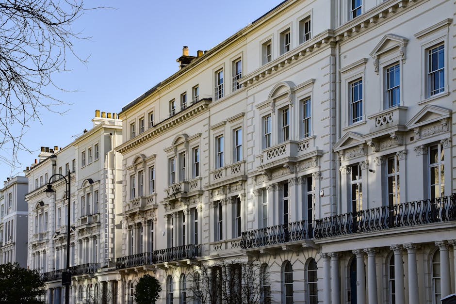 A row of historic white terraced townhouses with ornate architectural details, including decorative cornices, arched windows, and wrought iron balconies, set against a clear blue sky. The buildings feature a uniform facade with tall windows, some of which have small ledges or balustrades. The street level shows a paved sidewalk with a few small, neatly trimmed trees planted along the front, and a traditional black street lamp is visible on the left side of the image. Shadows cast by sunlight highlight the intricate stucco work and molding details on the facades of the buildings. The scene captures a quiet residential area in central London, characteristic of classic Georgian or Victorian architecture, offering an elegant backdrop that might relate to private or independent property management, similar to services provided by House Clearance Westminster, focusing on the preservation and maintenance of historic London buildings, often requiring careful waste handling and rubbish removal approaches while respecting the architectural integrity.
